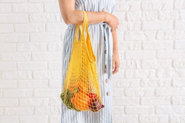 Woman Holding Mesh Eco Bag With Products Against White Brick Wal